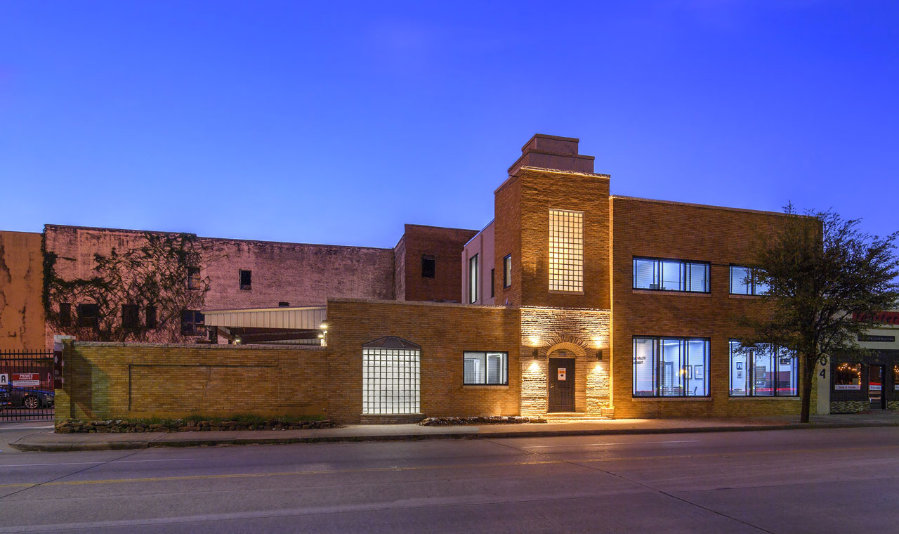 An evening shot of a stone office building with two floors and a spacious window, offering a glimpse of an office and a conference room. The entryway and frosted glass windows are illuminated.